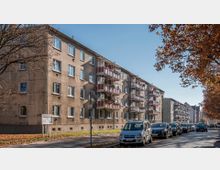 Wohngebäude mit vier Etagen und roten Balkonen an einer ruhigen Straße; am Straßenrand sind Autos geparkt, und im Vordergrund stehen Laubbäume mit herbstlichem Laub.