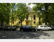 Gelbes Gebäude mit symmetrischer Fassade und hohen, bogenförmigen Fenstern, umgeben von Bäumen und einem Metallzaun. Im Vordergrund parkende Autos auf einer schattigen Straße.