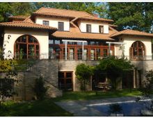 Villa mit mediterranem Stil, gekennzeichnet durch ein ziegelrotes Walmdach, große Holzfenster und runde Bögen. Vor der Fassade befindet sich ein Garten mit Terrasse, Kletterpflanzen und einem teilweise sichtbaren Pool.