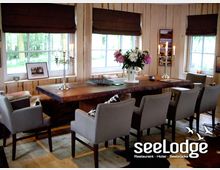 Gemütlicher Raum mit Holzwänden und großen Fenstern, eingerichtet mit einem rustikalen Holztisch, umgeben von grauen Polsterstühlen. Auf dem Tisch befinden sich Blumenvasen, Kerzenhalter und dekorative Elemente, während Tageslicht durch die Fenster einfällt.