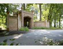 Backsteintorbau in neugotischem Stil mit spitzen Türmen und dekorativen Ornamenten, umgeben von Bäumen in einem parkähnlichen Umfeld. Eine Asphaltstraße führt durch das grüne Umfeld am Tor vorbei.