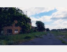 Verlassener Industriekomplex mit einem Backsteingebäude, das von Bäumen und Pflanzen teilweise überwuchert ist. Im Vordergrund eine asphaltierte, überwucherte Fläche, im Hintergrund weitere Gebäude und eine weitläufige Landschaft unter bewölktem Himmel.