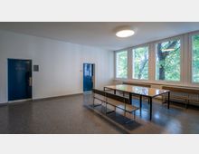 Ein schlichter Besprechungsraum mit einem rechteckigen Holztisch und Sitzbänken. Rechts befinden sich große Fenster mit Blick auf grüne Bäume, links ist eine blaue Tür mit der Beschriftung „Sitzungssaal“.