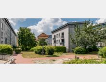 Wohngebiet mit mehreren mehrstöckigen Häusern, umgeben von gepflegten Grünflächen, Büschen und Bäumen. Im Vordergrund führt ein gepflasterter Weg durch den Innenhof, ein Holzüberdachungselement ragt in die Szene.