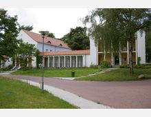 Weißer Gebäudekomplex mit roten Ziegeldächern, großen Fenstern und einer Kolonnade, umgeben von Grünflächen, Gehwegen und vereinzelt stehenden Bäumen. Im Vordergrund führt ein gepflasterter Weg durch die Anlage, während eine Straßenlaterne und eine Mülltonne sichtbar sind.