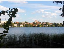 Blick auf eine Stadt am Ufer eines Sees, umgeben von Bäumen und Schilf im Vordergrund. Die Stadt besteht aus kleinen, teils historischen Gebäuden mit roten Dächern, darunter eine Kirche mit Turm in der Mitte, vor einem leicht bewölkten Himmel.