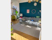 Wohnzimmer mit einer dunkelblauen Akzentwand, auf der einfache Wanddekorationen hängen. Im Vordergrund steht ein graues Sofa mit bunten Kissen, flankiert von kleinen Tischen mit Lämpchen und Accessoires. Der Raum hat einen Holzboden im Fischgrätenmuster, helle Teppiche, viele Zimmerpflanzen und eine Bücherwand neben einem Fenster mit viel Tageslicht.