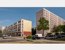 Mehrstöckige Wohnhäuser in einem städtischen Wohngebiet. Links ein langgestrecktes Gebäude mit Balkonen, rechts ein höheres Gebäude aus hellbeigen Fassadenelementen; davor parkende Autos, Gehwege und Bäume.