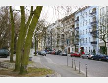 Straßenszene in einer städtischen Wohngegend mit mehrstöckigen Altbauten, teilweise renoviert und in hellen Farben gestrichen, dahinter reihenweise geparkte Autos. Im Vordergrund stehen Bäume und ein gepflasterter Gehweg mit niedrigen Begrenzungsmauern.