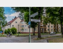 Straßenecke mit Wohngebäuden, umgeben von Bäumen und Grünflächen. Straßenschilder für „Haderslebener Straße“ und „Grillparzerstraße“ sind an einer Laterne befestigt; im Vordergrund fährt eine Person auf einem Fahrrad entlang einer gepflasterten Straße.