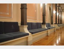 Innenansicht eines historischen Saals mit einer langen Reihe gepolsterter Sitzbänke in Blau, die durch dunkelbraune Säulen getrennt sind. Die Wände sind mit dekorativen Stuckelementen in orangefarbenem und weißem Putz gestaltet, der Boden besteht aus glänzend poliertem Holz.