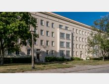 Mehrstöckiges Wohngebäude im Stil der Nachkriegsarchitektur mit hellem Putz und symmetrischen Fensterreihen, umgeben von Grünflächen, Bäumen und einem Gehweg im Vordergrund.