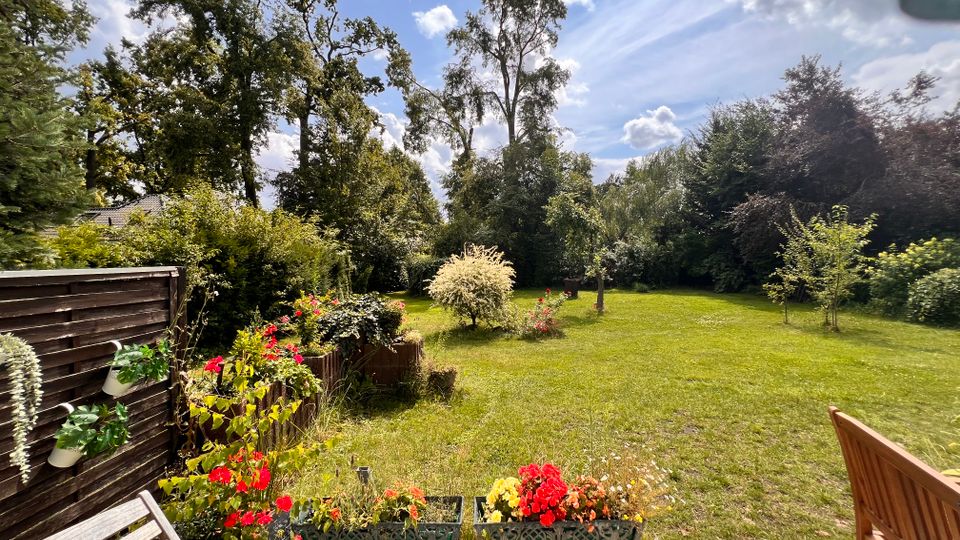 Blick von einer gefliesten Terrasse in einen großen Garten mit weitläufiger Rasenfläche, Blumen- und Pflanzkübeln im Vordergrund sowie einem Holzzaun links. Im Hintergrund stehen hohe Bäume und dichtes Buschwerk unter einem teils bewölkten Himmel.
