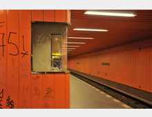 U-Bahn-Station mit orange verkleideten Wänden, durch Graffiti gekennzeichnet. Links ein gläserner Schaukasten, rechts die Gleise und der Bahnsteig mit der Aufschrift „Halemweg“.