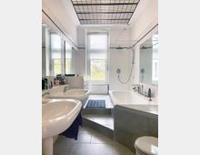 Helles Badezimmer mit zwei Waschbecken, einer Badewanne und einer ebenerdigen Dusche. Der Raum ist mit weißen Fliesen verkleidet, hat ein Fenster mit Tageslicht und eine Deckenstruktur mit schwarzen Gittermustern.