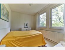 Schlicht eingerichtetes Schlafzimmer mit weiß gestrichenen Holzpaneelwänden, einem Bett mit gelbem Überwurf, einer Kommode und zwei großen Fenstern, die einen Blick ins Grüne bieten. An der Wand hängt eine Landkarte in einem blauen Rahmen.