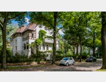 Weiße historische Villa mit Fachwerkelementen und rotem Ziegeldach, umgeben von Bäumen und dichtem Grün. Vor dem Gebäude führt eine gepflasterte Straße entlang, an der mehrere Autos parken.