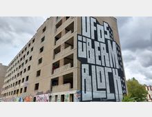 Verlassenes mehrstöckiges Gebäude mit offenstehenden Fensteröffnungen und teils beschmierten Wänden, auf dessen Fassade großformatige Graffiti-Kunst mit dem Schriftzug „UFOS ÜBERM BLOCK“ zu sehen ist. Im Vordergrund befinden sich weitere Graffitis sowie vereinzelte Bäume im Hintergrund unter bewölktem Himmel.