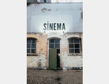 Außenansicht eines alten Backsteingebäudes mit großen Rundbogenfenstern und abgeblätterter Fassade. Über einer offenen, grün lackierten Tür ist in großen Buchstaben "SİNEMA" aufgemalt.