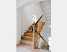 Holztreppe in einem hellen Innenraum mit weiß gestrichenen Wänden, einem Geländer aus Holz und schwarz lackiertem Handlauf. Im Hintergrund sind kleine Fenster mit natürlichem Lichteinfall sichtbar.