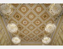 Decke eines prunkvollen historischen Saals mit reich verzierten Kassettendecken, zahlreichen farbigen Gemälden in den Feldern und goldenen Details. Mehrere große Kristalllüster hängen von der Decke, ergänzt durch dekorative Säulen und Wandleisten.