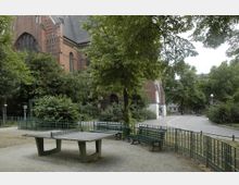 Backstein-Kirche in parkähnlicher Umgebung mit dichtem Baumbestand. Im Vordergrund stehen eine Tischtennisplatte sowie grüne Bänke und Zäune auf gepflastertem Boden.