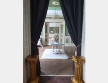 Blick in einen eleganten Salon mit grün tapezierten Wänden, einem Kronleuchter und einem verzierten Tisch auf einem Teppich. Der Raum ist durch schwarze Vorhänge mit goldfarbenen Säulen am Eingang eingerahmt.