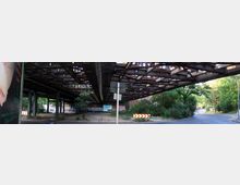 Eine Straßenunterführung mit einer rostigen Stahlkonstruktion darüber, umgeben von Pflanzen und Bäumen. Im Hintergrund sind Graffitis, ein Parkplatz und ein Gehweg sichtbar.