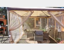 Überdachter Gartenpavillon mit beigefarbenem Stoffdach und transparenten Seitenwänden, innen ein Tisch mit mehreren Rattan-Stühlen auf einem runden Teppich. Links steht ein gemauerter Grillkamin auf der Terrasse, im Hintergrund sind Bäume und Gartenbewuchs zu sehen.