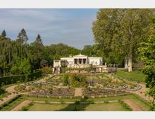 Historisches Gebäude im klassizistischen Stil, umgeben von einem symmetrischen Garten mit gekrümmten Wegen und Blumenbeeten. Im Hintergrund dichter Wald, während hohe Bäume und Hecken den Garten einrahmen.