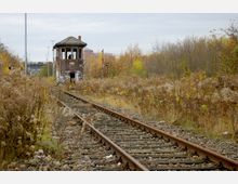 Ehemalige Bahngleise führen zu einem kleinen, verlassenen Stellwerkgebäude aus Backstein, das mit Graffiti bedeckt ist. Das Gelände ist von verwilderter Vegetation und herbstlich gefärbten Bäumen umgeben.