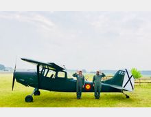 Zwei Personen in grünen Fliegeranzügen salutieren vor einem dunkelgrünen historischen Propellerflugzeug auf einer Wiese. Im Hintergrund sind ein Holzzaun, Felder und Wald zu sehen.