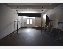 Ein schlichter, rechteckiger Raum mit weiß gestrichenen Wänden und einer Kombination aus dunklem Boden und gemusterten Fliesen. Zu den sichtbaren Elementen gehören ein Kleiderständer, Licht- und Fotoausrüstung sowie Möbel wie ein kleiner runder Tisch, eine Bank und Stühle; im Hintergrund eine Doppeltür mit Milchglasfenstern.