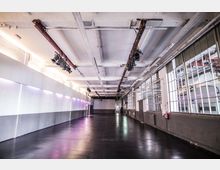 Langer, rechteckiger Veranstaltungsraum mit industriellem Charakter, hohen Decken und sichtbaren Beleuchtungseinrichtungen. Auf der linken Seite eine glatte Wand mit violetter Beleuchtung, auf der rechten Seite große Fenster mit Metallrahmen, die viel Licht hereinlassen.