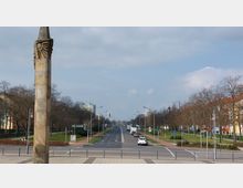 Eine breite Straße mit vier Fahrspuren, flankiert von Fuß- und Radwegen sowie bepflanzten Grünflächen mit kahlen Bäumen. Links und rechts der Straße sind Wohngebäude und Laternen zu sehen, im Vordergrund steht eine steinerne Säule mit Verzierungen.