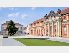 Hauptmotiv ist das Filmmuseum Potsdam, ein barocker Bau mit einer roten Fassade, weißen Fensterrahmen, dekorativen Säulen und Skulpturen an der Front. Im Vordergrund sind eine Grünfläche, ein gepflasterter Platz und ein Schild mit der Aufschrift „Filmmuseum Kino“ zu sehen.