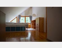 Dachzimmer mit schrägem, weiß gestrichenem Dach, einem Holzboden und einem großen Fenster mit hellblauen Vorhängen, das auf eine grüne Waldlandschaft blickt. Der Raum ist mit einem Bett, einer Holzkommode und einem offenen Regal aus hellem Holz möbliert.