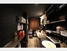 Schmaler Küchen- und Vorbereitungsraum mit dunklen Wänden, Fliesenboden und Arbeitsfläche aus Stein, flankiert von Regalen mit Behältern, Küchenutensilien und Gewürzflaschen. Am Ende des Raums steht ein großer Holzschrank unter Deckenbeleuchtung.