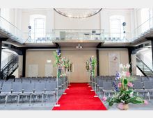 Ein moderner Veranstaltungssaal mit einer roten Teppichbahn in der Mitte, flankiert von mit Blumen dekorierten Ständern. Im Hintergrund befinden sich graue Stuhlreihen, eine Empore mit Glasgeländer und eine Deckenleuchte mit hängenden Glaskugeln.