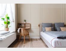 Helles Schlafzimmer mit Holzboden, einem Doppelbett mit blauen Kissen und beigen Akzenten, einer Fensterbank mit Sitzkissen und einer großen grünen Zimmerpflanze. Daneben steht ein kleiner Schreibtisch mit einem geflochtenen Holzstuhl und dekorativen Pflanzen.