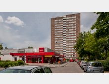 Hochhaus aus rotem Backstein im Hintergrund, davor ein flaches, modernes Gebäude eines Penny-Supermarktes mit roter Fassade und großen Fenstern. Umgeben von parkenden Autos, Bäumen und einer Straßenszene an der Auersbergstraße.