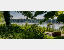 Garten am Ufer eines Sees mit dichten Büschen und Bäumen im Vordergrund, einem Holzdeck mit Liegestuhl und einer begrünten Metallpergola. Im Hintergrund befindet sich der See mit Booten und einer bewaldeten Küstenlinie.