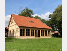 Fachwerkhaus mit rotem Satteldach und großen, bodentiefen Fenstern, umgeben von einer Wiese und vereinzelten Bäumen. Das Gebäude steht in einer ländlichen Umgebung bei blauem Himmel.