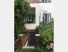 Außenansicht eines Hauses mit weiß verputzter Fassade und roten Klinkerelementen. Im Vordergrund befinden sich eine Treppe mit einem Gittertor, umgeben von dichter Vegetation, einschließlich Bambus und blühenden Pflanzen, sowie eine Terrasse mit Fenstern und einem Sonnenschirm.