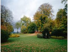 Herbstlicher Park mit weiten Rasenflächen, Laubbäumen in gelb-grünen Farbtönen und einer kleinen Statue auf einem Sockel in der Mitte. Im Hintergrund ist ein schmaler Weg sowie eine offene Landschaft mit Blick auf einen See erkennbar.