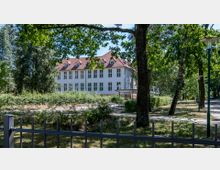 Ein zweistöckiges, historisches Gebäude mit weißer Fassade und rotem Ziegeldach, umgeben von Bäumen und Sträuchern. Im Vordergrund sind ein Zaun und ein gepflasterter Gehweg zu sehen, die durch einen parkähnlichen Bereich führen.