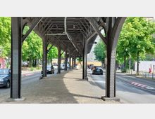 Straßenansicht unter einer erhöhten Eisenbahntrasse mit Metallstützen und Gitterstruktur. Auf beiden Seiten der Straße stehen Bäume, und Autos fahren in beide Richtungen.