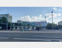 Straßenansicht des Restaurants "Moskau" in Berlin, einem modernen Gebäude mit breiten Glasfronten im oberen Stockwerk, einer schlichten Fassade im Erdgeschoss und dem Schriftzug "MOSKAU" auf dem Dach. Ein Werbeplakat und vereinzelte Menschen sowie geparkte Fahrräder und Autos sind im Vordergrund sichtbar.