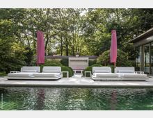 Moderner Außenbereich mit Pool, umgeben von Bäumen und Sträuchern. Vier weiße Sonnenliegen mit rosa Sonnenschirmen stehen auf einer gepflasterten Fläche vor einem gemauerten Kamin und gepflegter Gartenlandschaft.