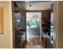 Moderne Küche mit Holzboden, grauen Hochglanzschränken und einer offenen Ansicht zu einem lichtdurchfluteten Essbereich mit großen Fenstern und Blick auf den Garten. Der Raum ist mit diversen Küchenutensilien und Dekorationselementen ausgestattet.